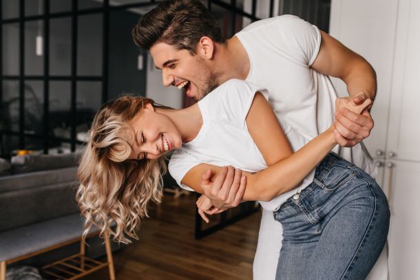 Wonderful girl in white t-shirt dancing with boyfriend. Indoor photo of winsome lady fooling around at home with husband.